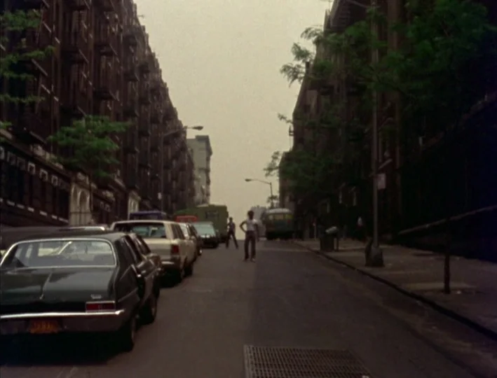 News From Home (1977) movie still: wide — A narrow city street is flanked by tall, brown brick apartment buildings with fire escapes. A young person…