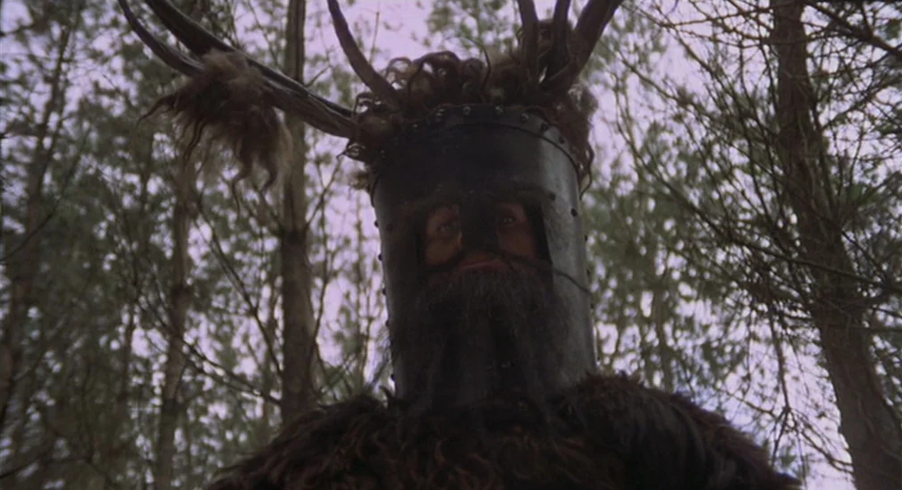 Monty Python and the Holy Grail (1975) movie still: closeup — A person wears a large metal helmet topped with antlers inside a dense forest. The…