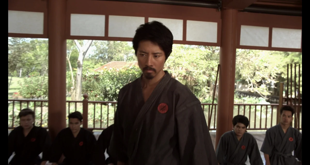 Ninja: Shadow of a Tear (2013) movie still: medium — A wooden dojo structure with open walls looks out onto a leafy garden. A man in a dark martial…