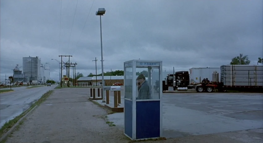 About Schmidt movie still: wide — A concrete lot features a solitary blue telephone booth under a gray sky. A man in a flat cap stands inside the…