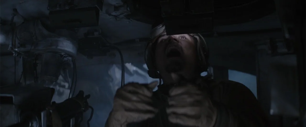 Fury (2014) movie still: medium — A cramped, dimly lit steel interior filled with mechanical levers and communications equipment. A man with a…