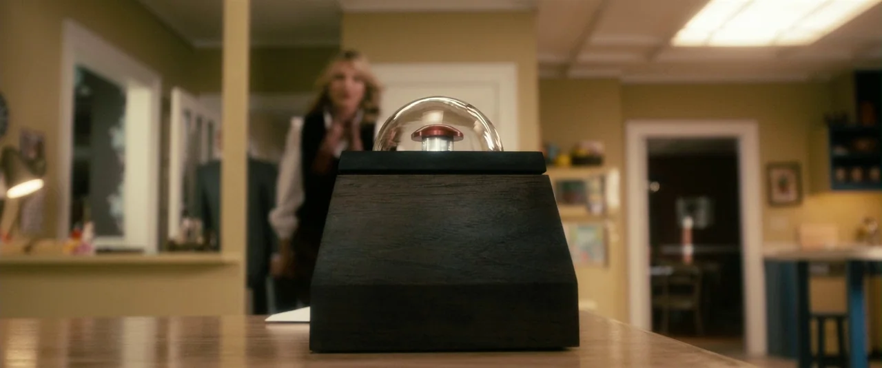 The Box (2009) movie still: closeup — A wooden box containing a large red button under a glass dome sits in focus. In the blurred background, a…
