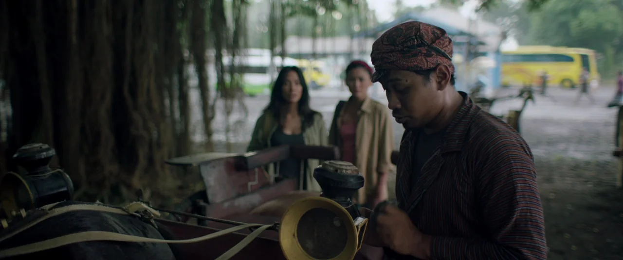Impetigore (2019) movie still: medium — An outdoor transport area features a large banyan tree with hanging roots overhead. A man works on a horse…