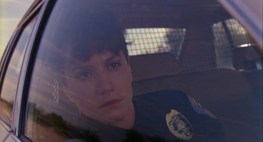Raising Arizona (1987) movie still: medium — A view from outside a police vehicle looking through the side glass window. A young female officer sits…