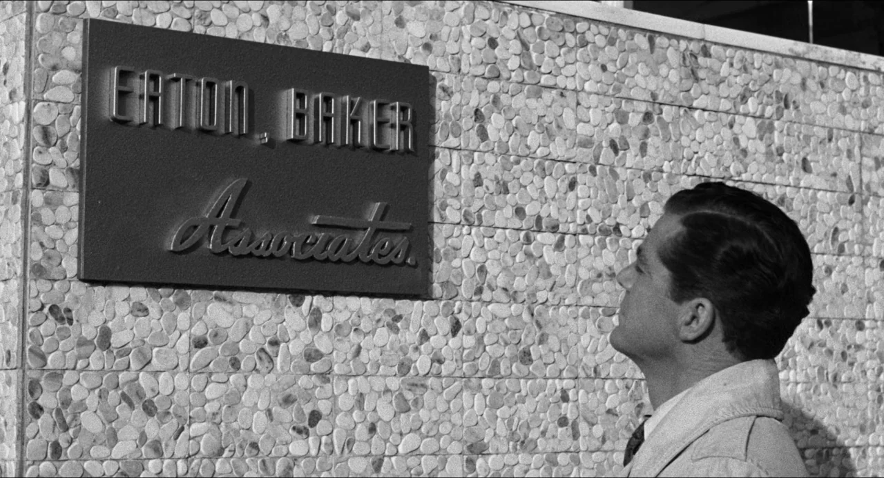 The Fearmakers (1958) movie still: medium — An exterior wall made of grey river stones features a rectangular company nameplate. A man in a light…