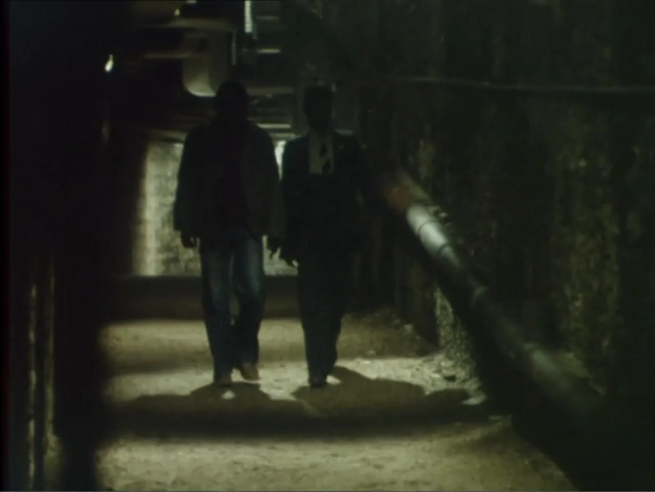 A Dessert for Constance (1981) movie still: fullbody — A dark, narrow underground passage features rough walls and pipes running along the side. Two…