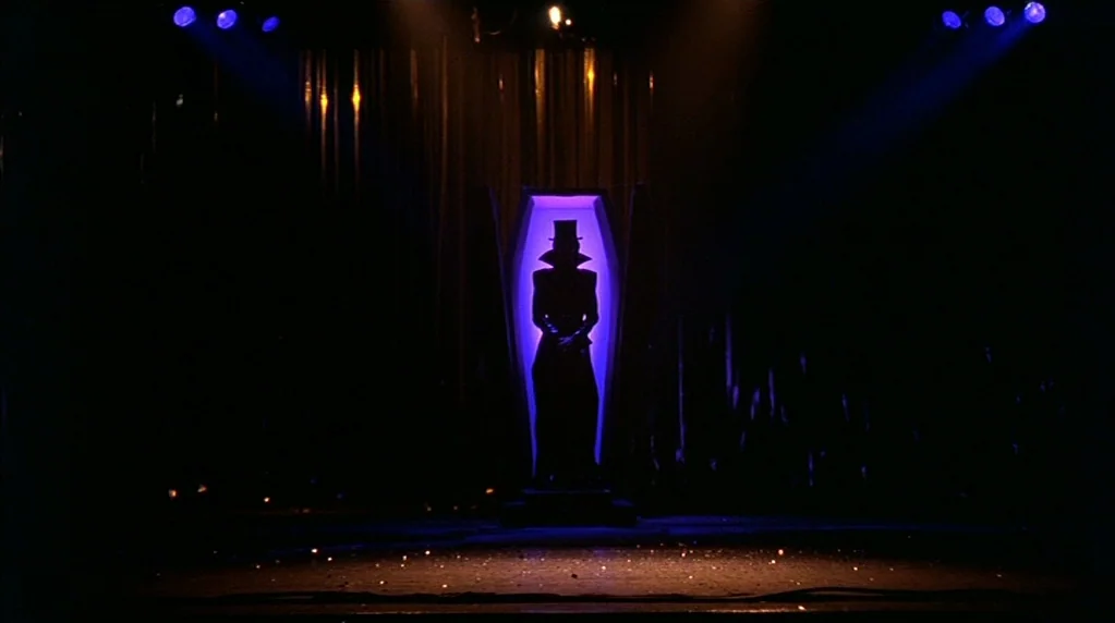 Velvet Goldmine (1998) movie still: wide — A dark stage with a central vertical cabinet containing a person in silhouette. The person stands…