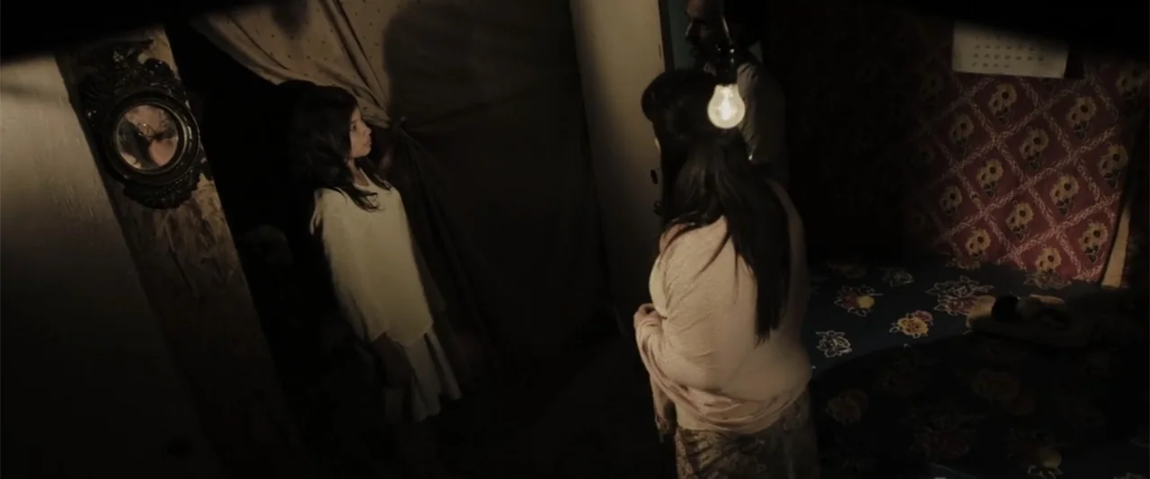 At Dawn (2013) movie still: medium — A dimly lit, cramped interior room features patterned fabric on the wall and a mirror. Two women stand facing…