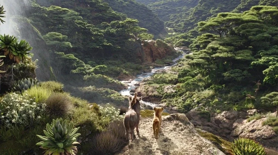 The Lion King (2019) movie still: wide — A vast lush valley features a flowing river surrounded by dense green vegetation. A warthog and a small…