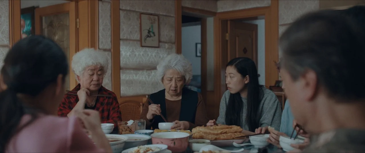 The Farewell (2019) movie still: medium — A group gathers at a dinner table set with several bowls and a platter. Individuals hold chopsticks while…
