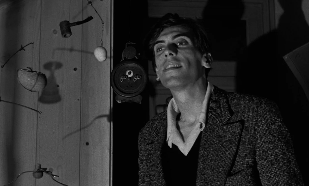 La Verite (1960) movie still: medium — A dimly lit room contains hanging abstract wire sculptures against a plain wooden wall. A young man wearing a…