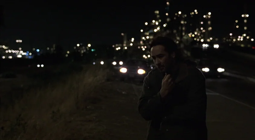 Being John Malkovich (1999) movie still: medium — A man stands on a roadside at night near an industrial area with distant lights. He rests his hand…