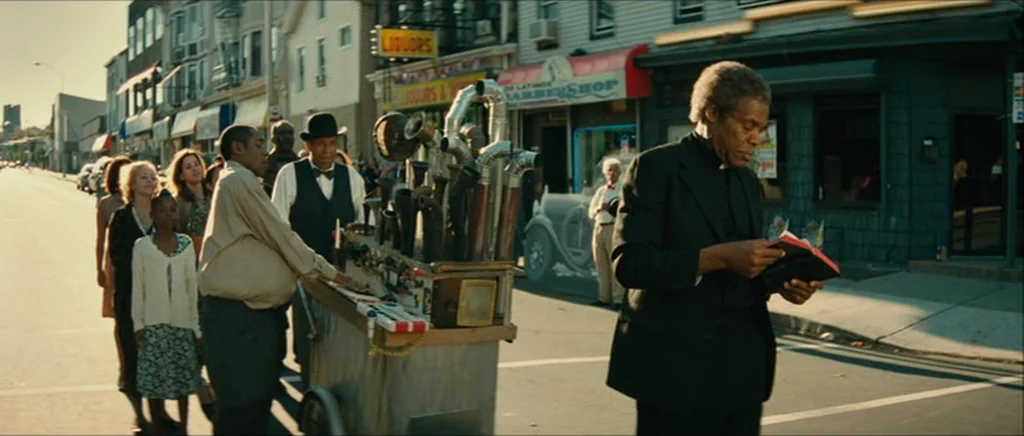 Be Kind Rewind (2008) movie still: wide — An urban intersection with businesses including a barbershop and a liquor store. An older man in a suit…