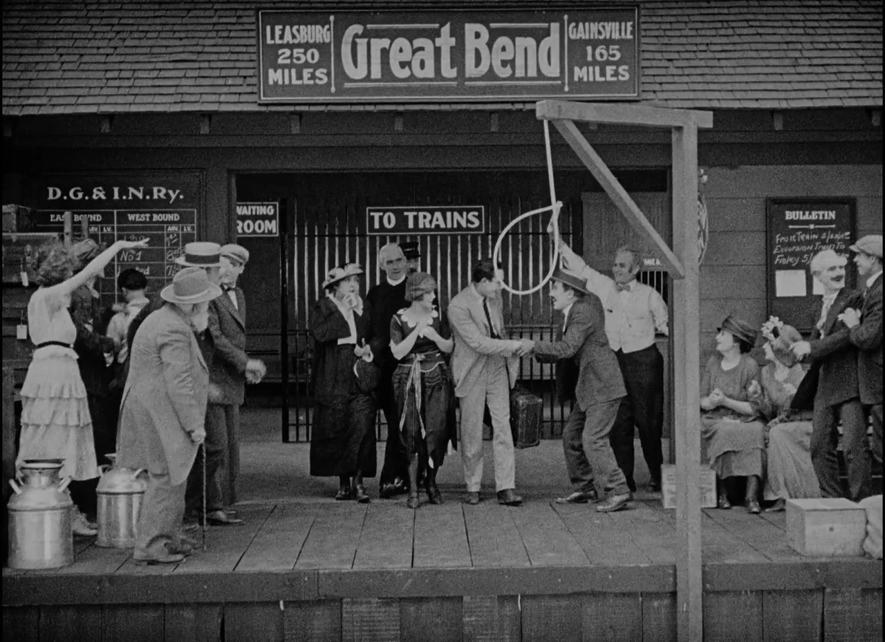 Safety Last! (1923) movie still: wide — A wooden train station platform features a prominent gallows-like post with a rope noose. A large group of…