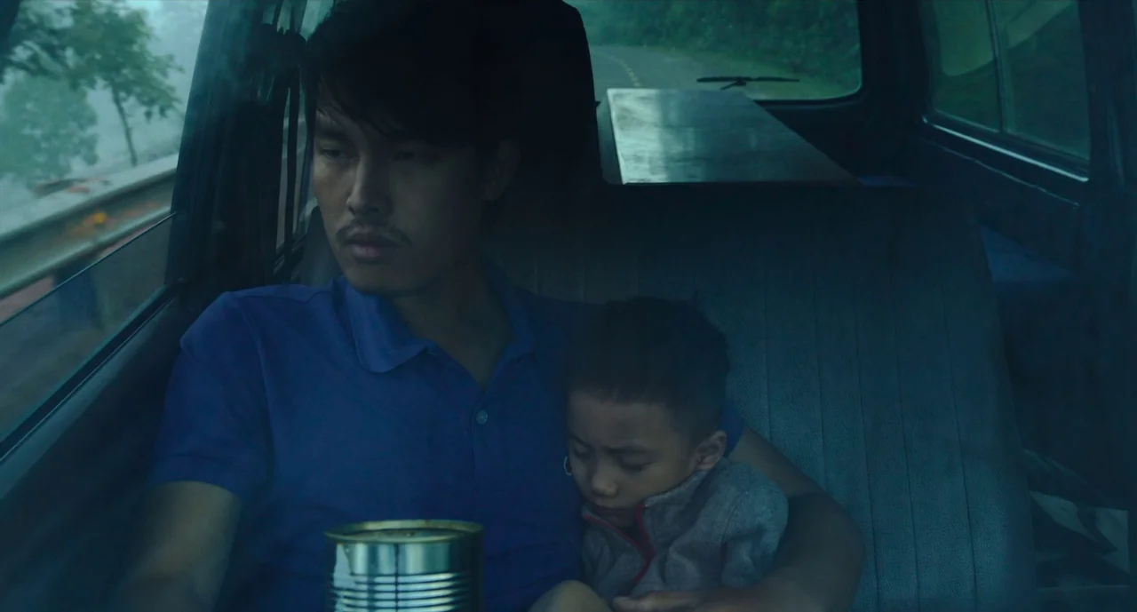 Inside The Yellow Cocoon Shell (2023) movie still: medium — The interior of a vehicle features a man sitting with a sleeping small child. Through…