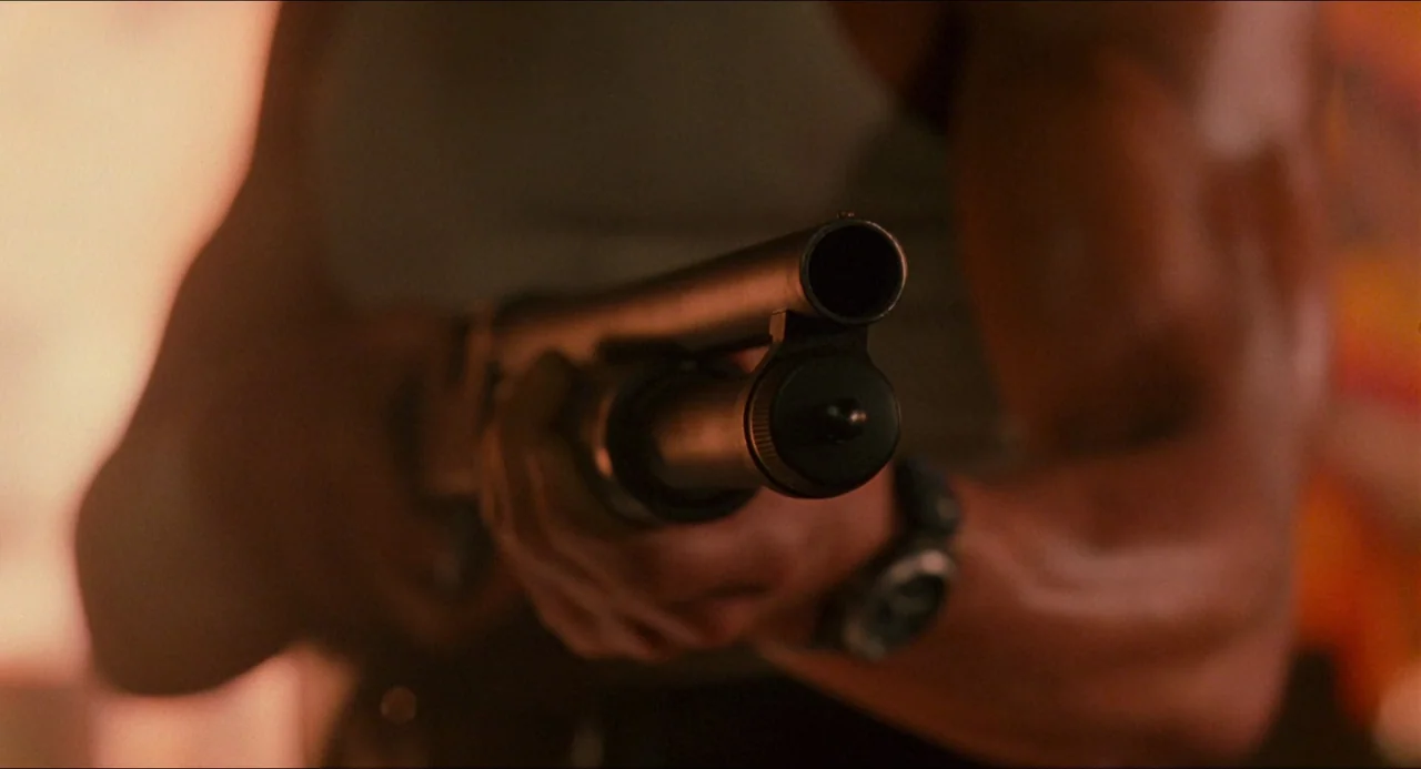 Hard Target (1993) movie still: closeup — A muscular man in a grey ribbed tank top holds a shotgun barrel forward. The weapon is aimed directly at…