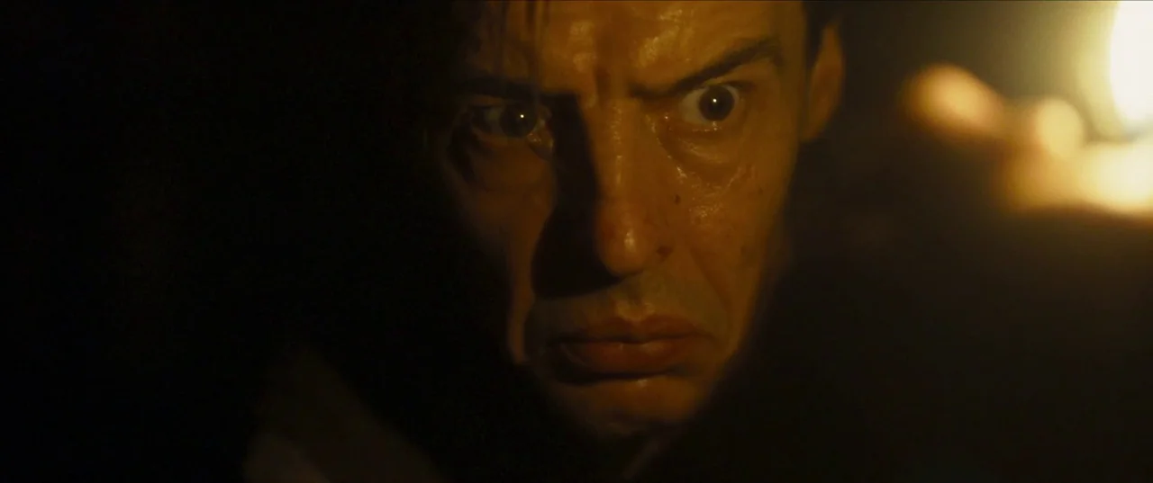 Earwig (2022) movie still: closeup — A dark, undifferentiated space provides the backdrop for a tight facial close-up shot. The man stares intensely…