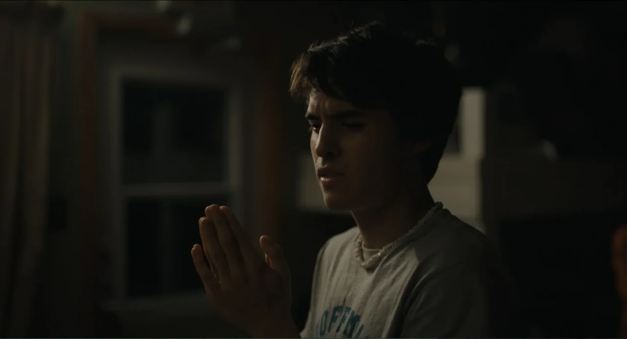 Hell of a Summer (2025) movie still: medium — A dimly lit residential room interior featuring a window frame in the dark background. A young male…