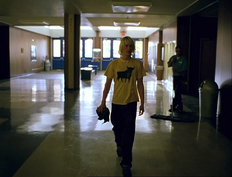 Elephant (2003) movie still: fullbody — A long, institutional hallway features polished linoleum floors and beige paneled walls. A young man walks…