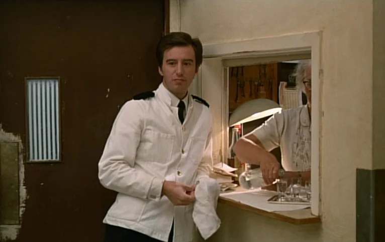 Giliap (1975) movie still: medium — A man in a white service uniform stands beside a wall-mounted hatch. Inside the hatch, an older woman pours…