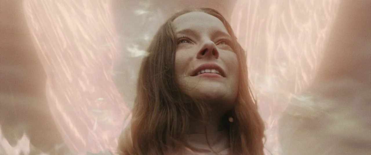Saint Maud (2019) movie still: closeup — A woman stands in a softly illuminated environment with shimmering light overlays. She gazes upward with a…