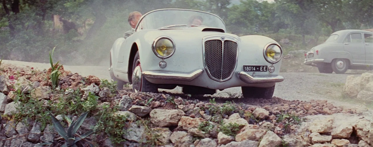 And God Created Woman (1956) movie still: medium — A cream-colored vintage Lancia convertible drives over a rocky, uneven dirt road. The vehicle…
