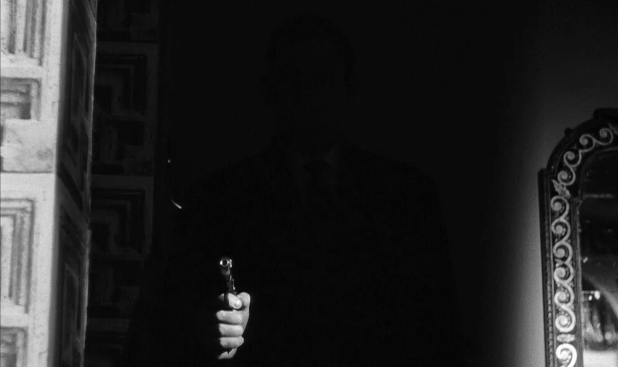 Le Doulos (1962) movie still: medium — A dimly lit, narrow corridor features patterned wallpaper on one side and an ornate mirror. A man stands in…