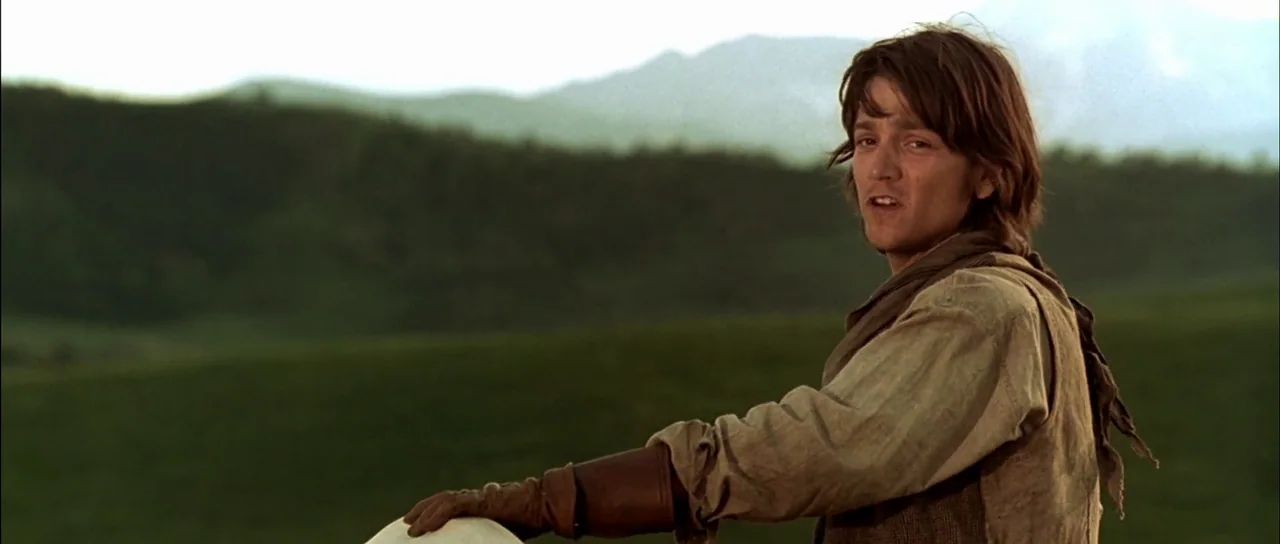 Open Range (2003) movie still: medium — A vast green landscape under a bright sky with distant rolling hills visible. A young man with brown hair…