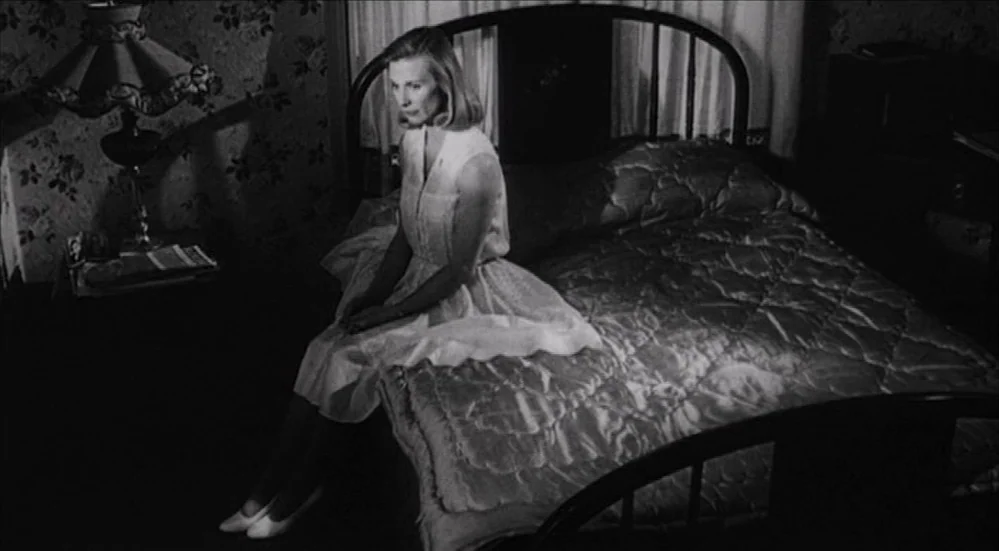 The Last Picture Show (1971) movie still: medium — A dimly lit room contains a double bed with a quilt and floral wallpaper. A woman in a light…