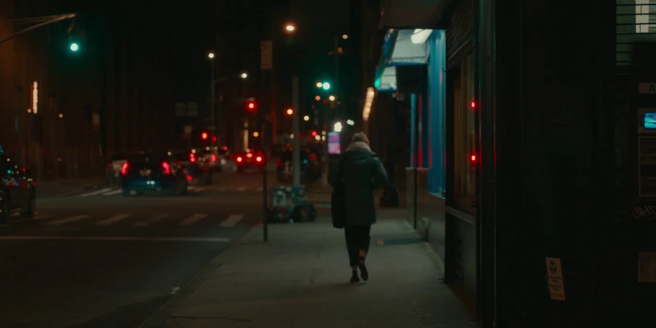The Assistant (2019) movie still: fullbody — A dark city sidewalk at night with a storefront facade on the right side. A lone pedestrian walks away…