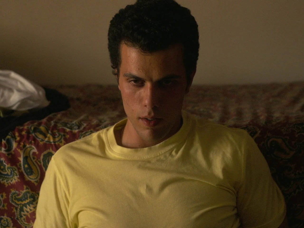 Of An Age (2022) movie still: medium — A dimly lit room features a patterned textile covering a bed surface. A man in a yellow shirt sits on the…