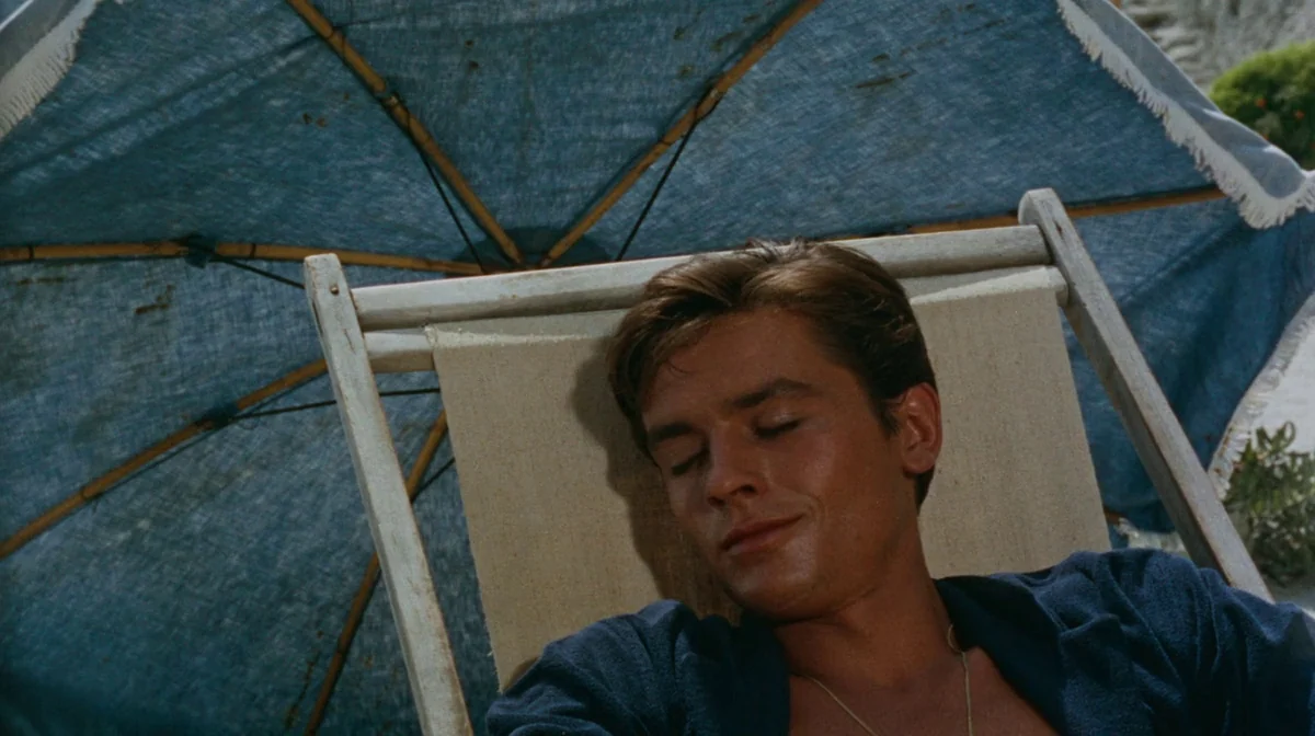 Purple Noon (Plein Soleil) (1960) movie still: closeup — A man rests on a wooden lounge chair beneath a blue sun umbrella. The beach setting is…