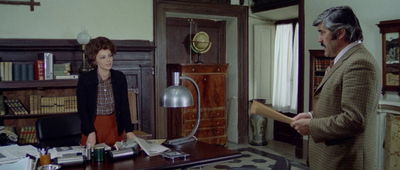 What Have They Done To Your Daughters? (1974) movie still: medium — A cluttered 1970s study with wood paneling and a large desk. A woman stands…