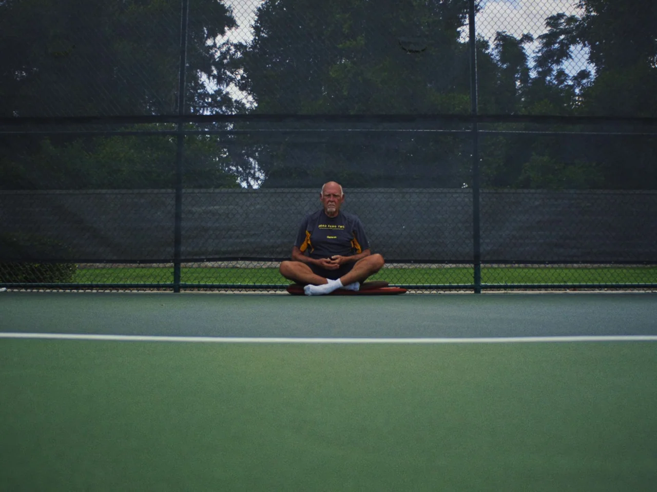 Some Kind Of Heaven (2020) movie still: fullbody — A man sits cross-legged on a green tennis court against a chain-link fence backdrop. The court…
