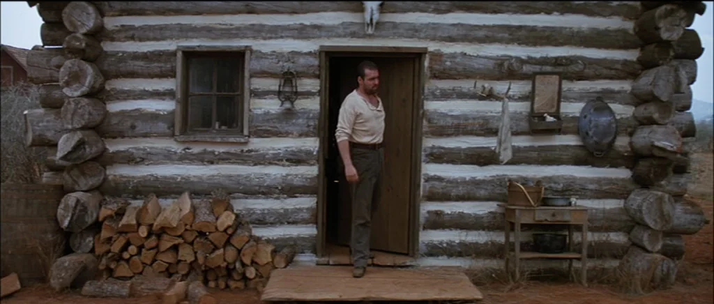 Unforgiven (1992) movie still: fullbody — A rustic log cabin exterior features white mortar between the horizontal dark timber logs. A man in a…
