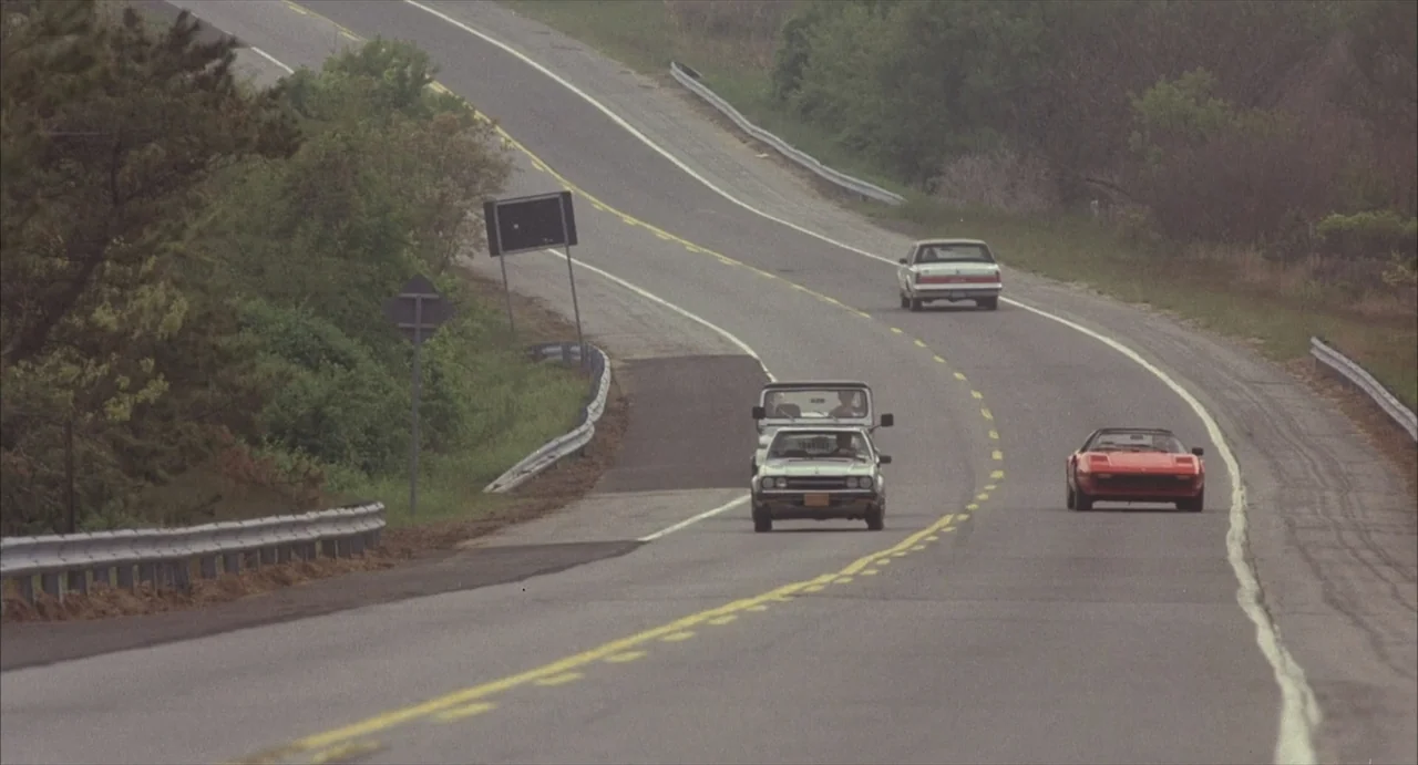Masquerade (2023) movie still: wide — An asphalt highway winds through a rural landscape with sparse trees and guardrails. Three vintage cars travel…