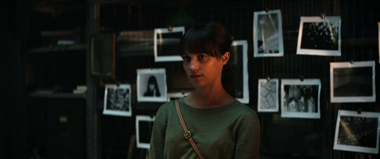 Earthquake Bird movie still: medium — A dark, cluttered space with numerous black and white photographs clipped to a wire rack. A woman stands in…