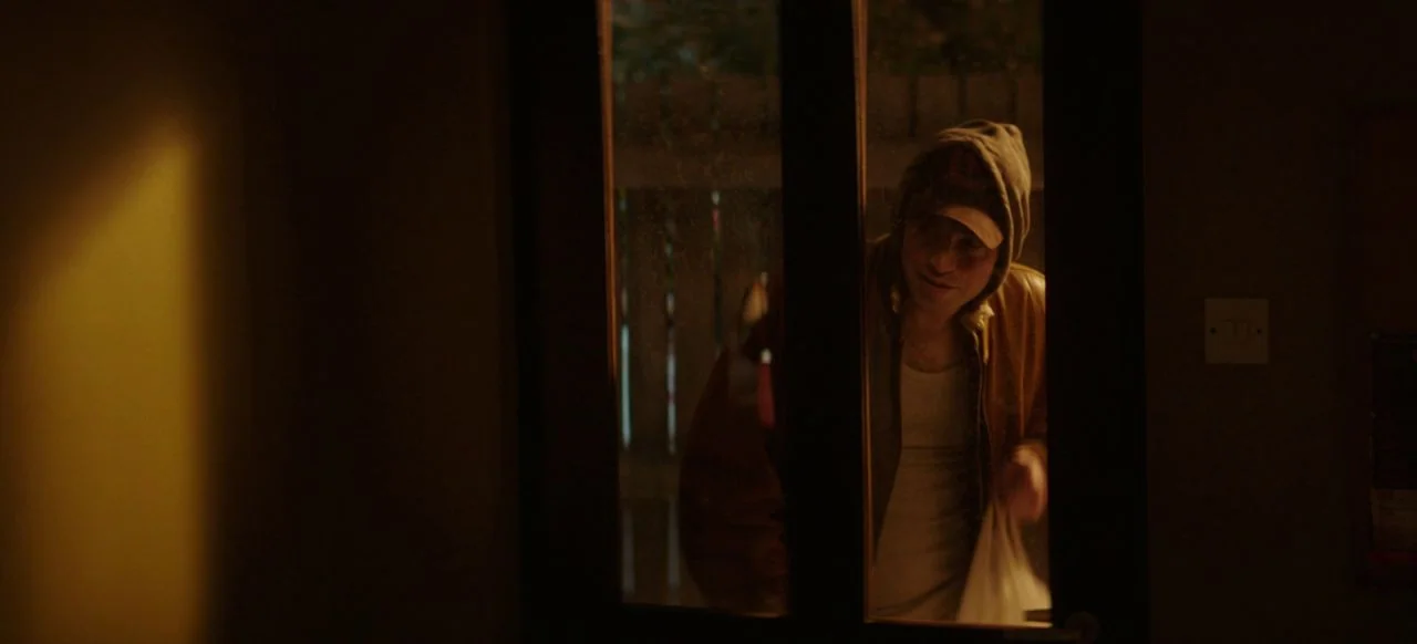A Good Woman Is Hard To Find (2019) movie still: medium — A man stands outside a glass-paneled door in a dark, warm-lit environment. He carries a…