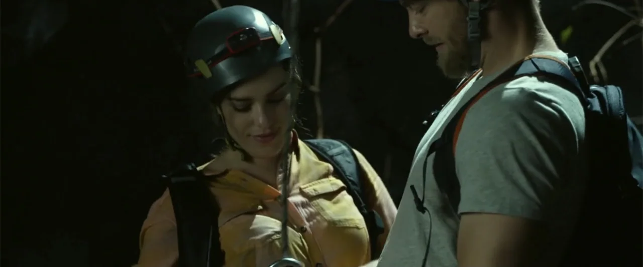 Black Water: Abyss (2020) movie still: medium — A dimly lit rocky environment with two people equipped for a cave exploration. The subjects face…