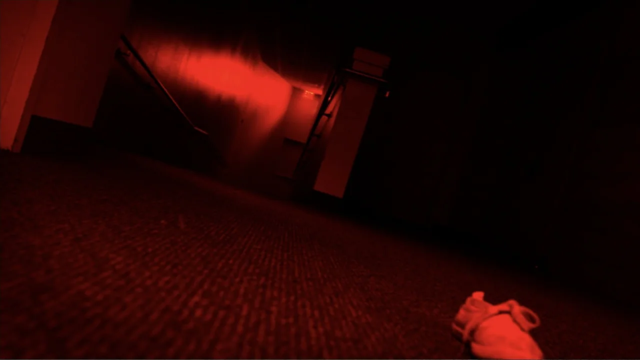 The Gallows (2015) movie still: establishing — A darkened interior hotel corridor features red emergency lighting and a carpeted floor. A single…