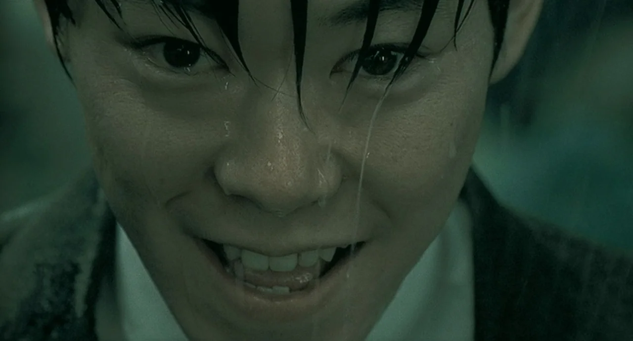 Uzumaki (2000) movie still: closeup — A close-up view of a person in the rain during a dark night. The individual stares directly at the viewer with…