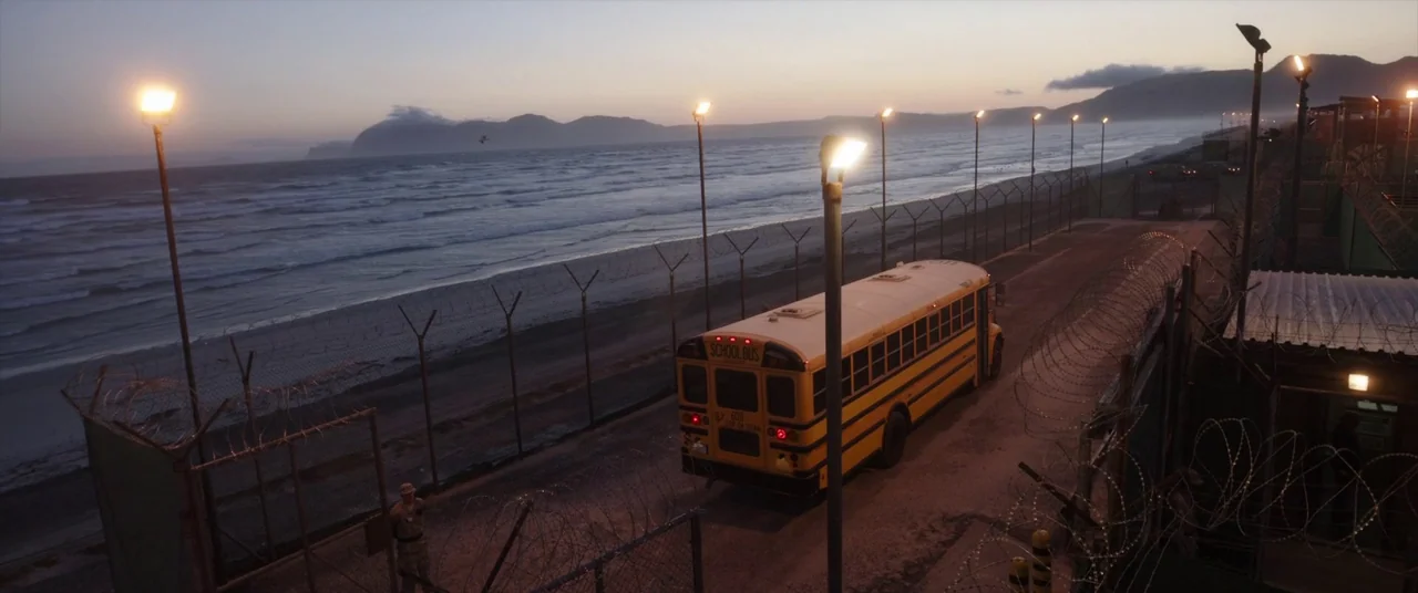The Mauritanian (2021) movie still: wide — A yellow school bus travels down a road lined with barbed wire fencing beside the ocean. Mountainous…