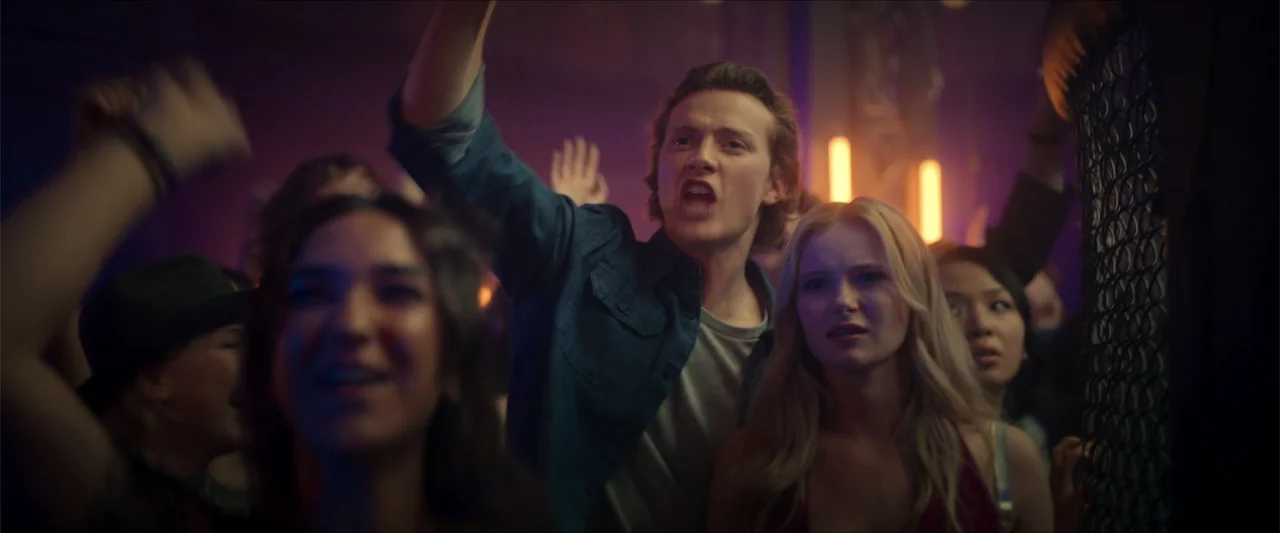 Love Me Love Me (2026) movie still: medium — A crowded, dimly lit nightclub interior filled with people moving in the dark. A young man and a blonde…