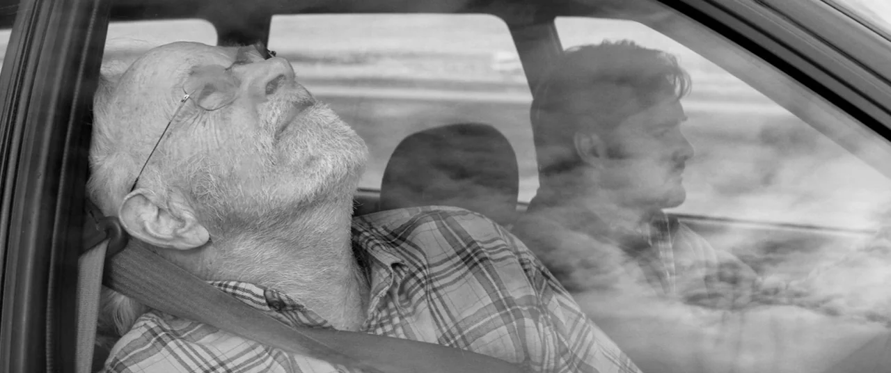 Nebraska (2013) movie still: medium — The interior of a moving car shows two men traveling down a highway. An elderly man sleeps in the passenger…