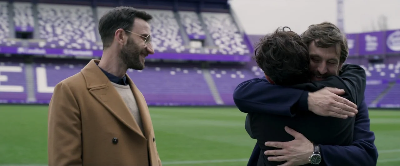 Voy a pasármelo bien (2022) movie still: medium — A professional soccer pitch features empty purple stadium seating in the background. Two men…