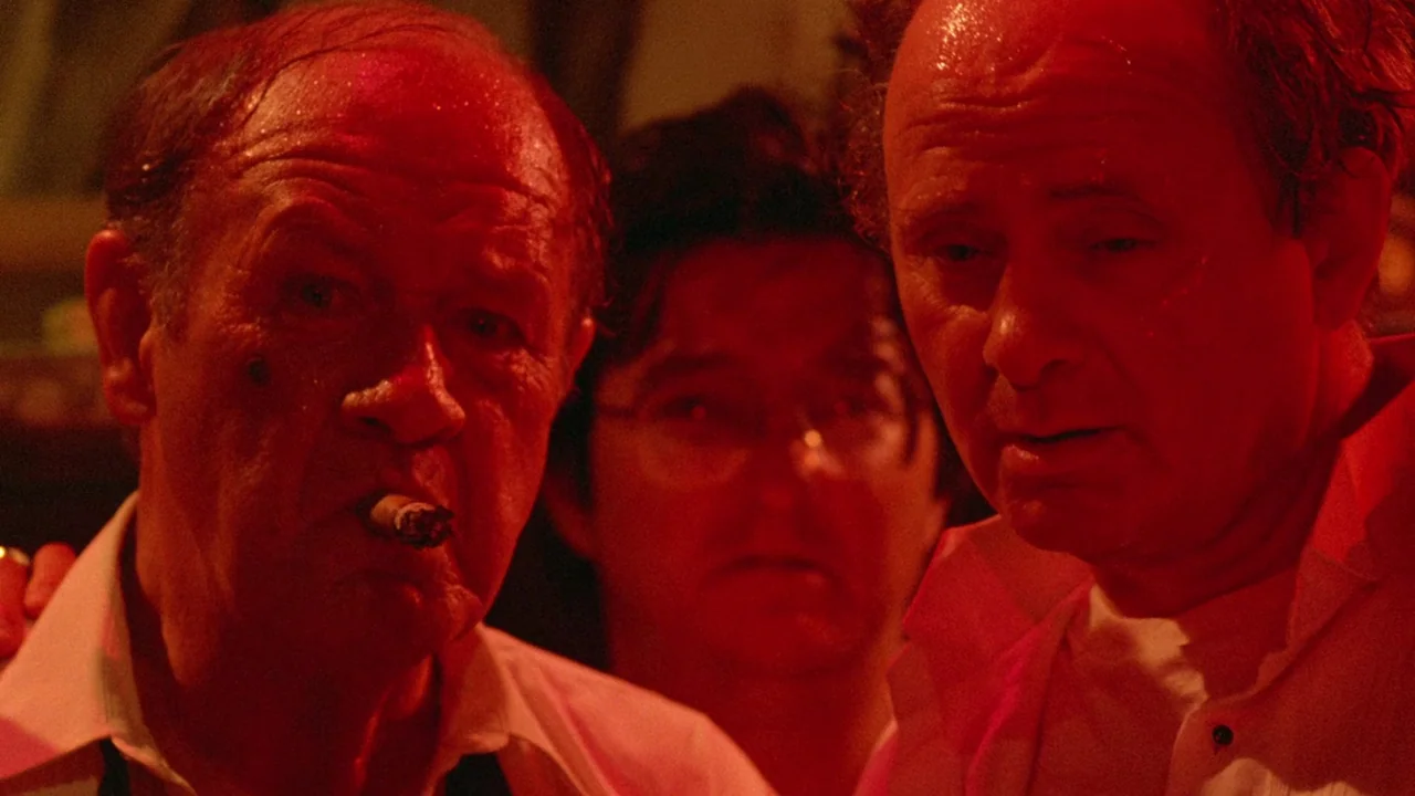 Society (1989) movie still: medium — A dimly lit room with heavy red light casting shadows on three men. The subjects stand close to the camera…