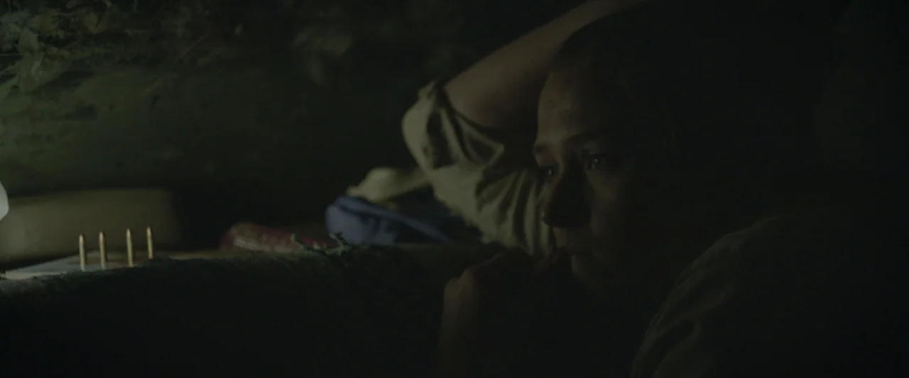 Here Alone movie still: closeup — A dark subterranean enclosure with earthen walls and minimal light. A young woman lies in the dim space with small…