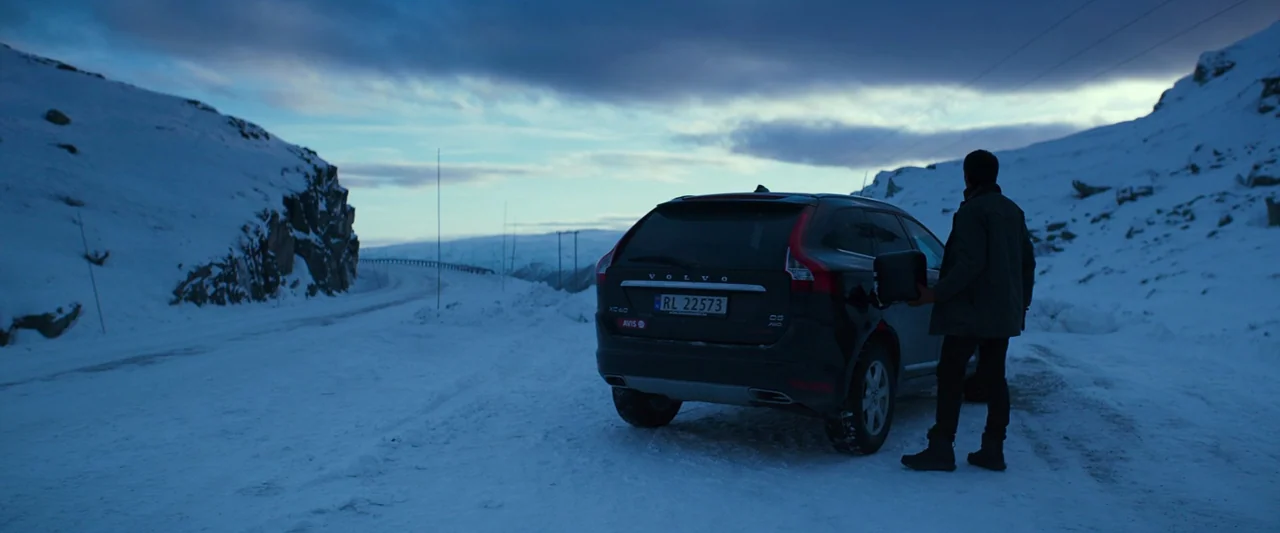 Believer (2018) movie still: wide — A dark SUV is parked on a snowy road between two steep, snow-covered mountains. A lone figure stands near the…