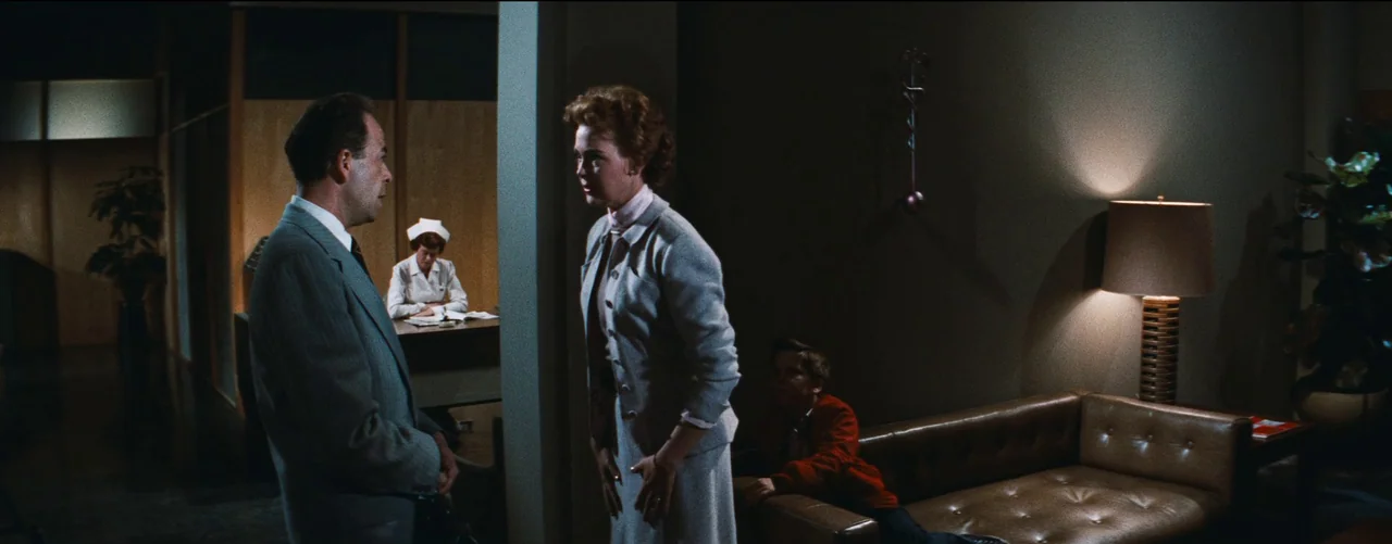 Bigger Than Life (1956) movie still: wide — A 1950s professional space with wood paneling and a tufted leather sofa. A man and woman converse while…