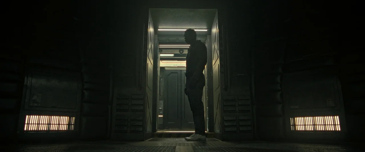 Alien: Romulus (2024) movie still: fullbody — A dark industrial corridor features a brightly lit doorway at the center. A man stands in silhouette,…